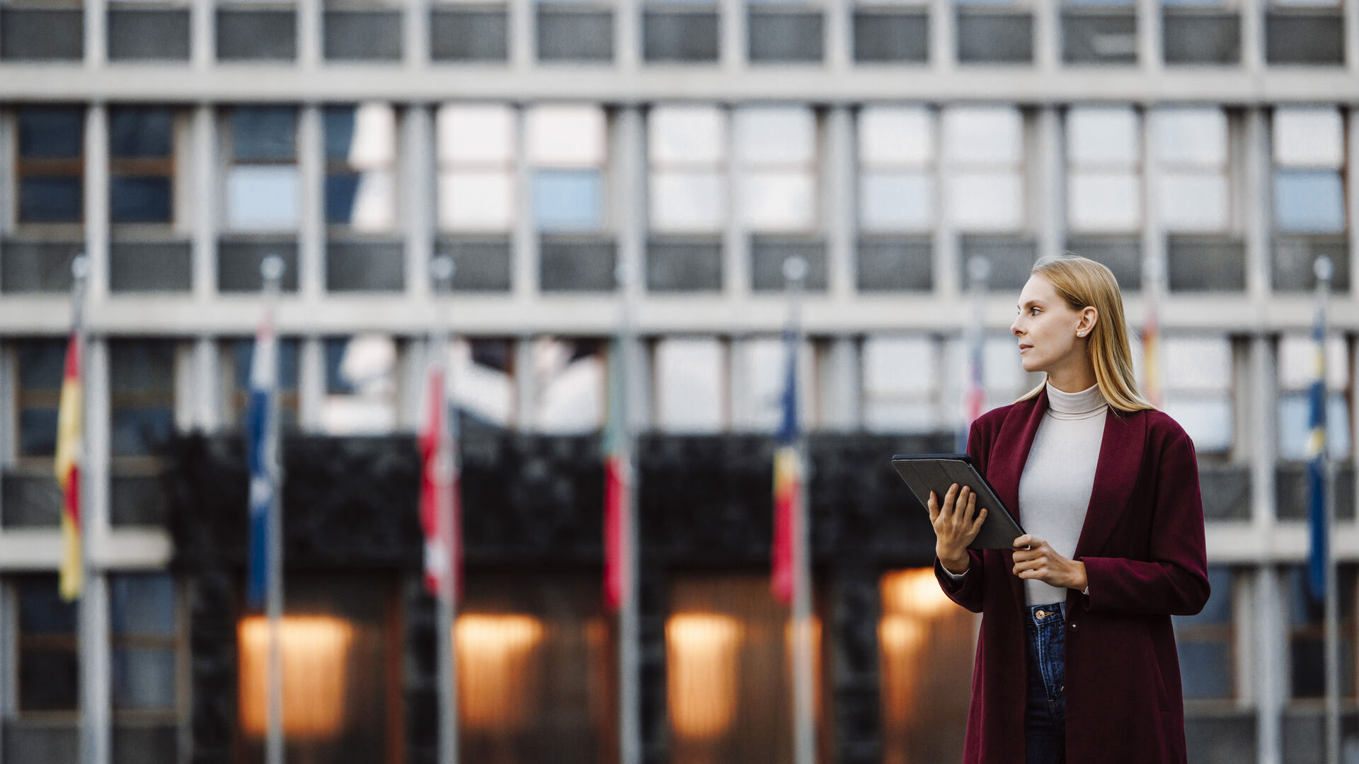 A young woman holding a tablet in front of a public building with flags